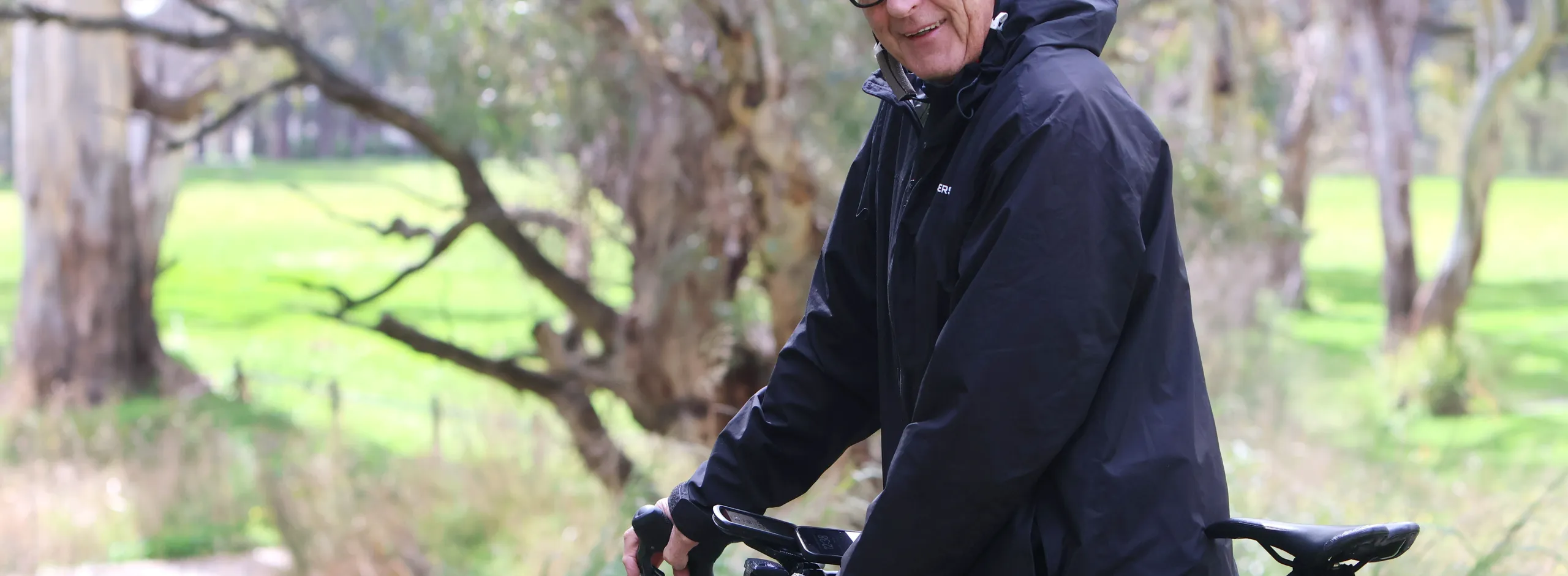 Mayor Bim Lange seated on his bike, wearing helmet and glasses, ready to go for the ride. Large gum trees are in the background