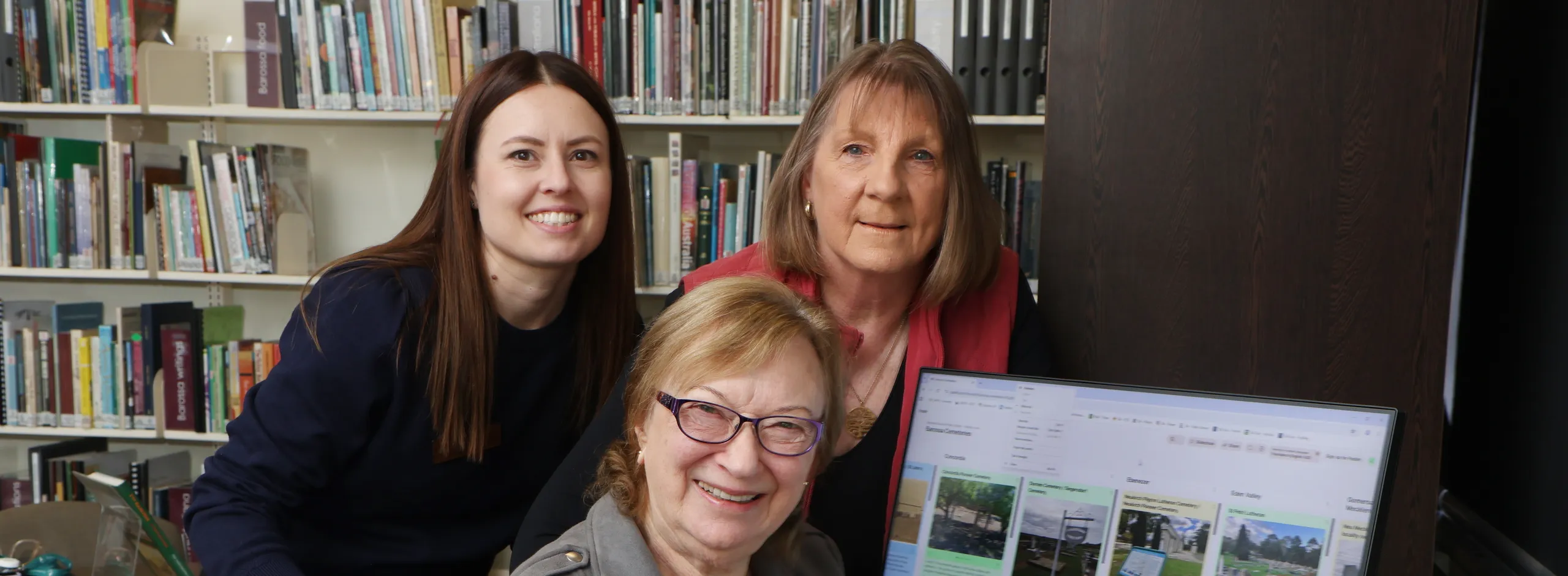 Three women seated around a computer screen which is showing the new online Barossa Cemeteries family research tool that goes live on 27 August.