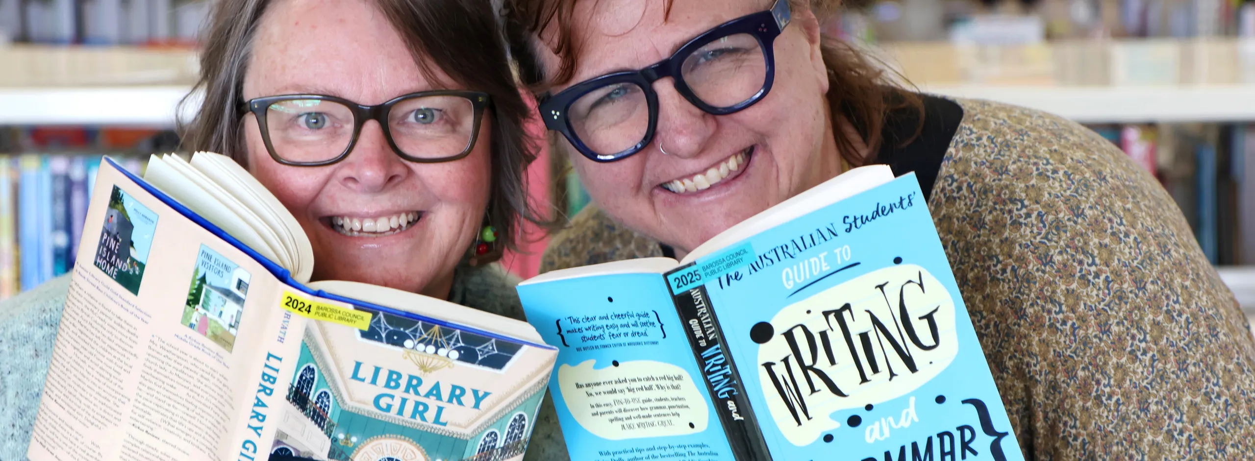 Two wmiling women peaking out from behind the books they are holding in front of book shelves in a library.