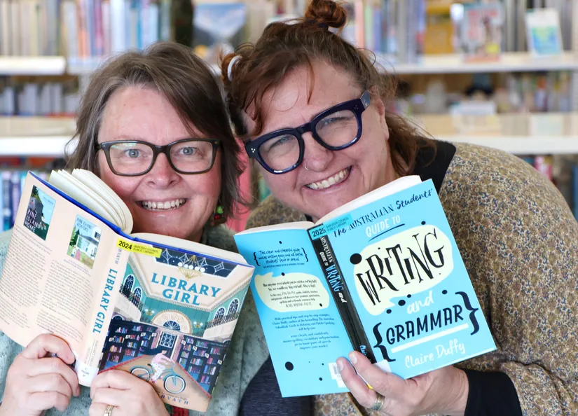 Two wmiling women peaking out from behind the books they are holding in front of book shelves in a library.