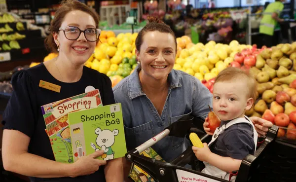Library staff member holding books, chatting to a mum and her son who is holding fruit and sitting in a shopping trolley.