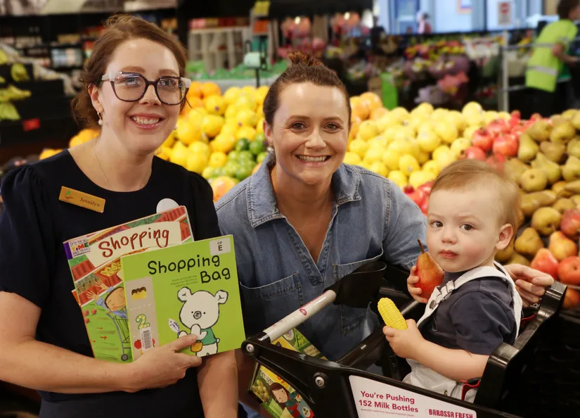 Library staff member holding books, chatting to a mum and her son who is holding fruit and sitting in a shopping trolley.