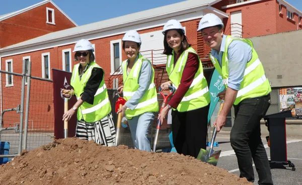 Four people wearing hard hats and holding shovels photographed in the carpark (with original red-brick hall behind), during the official sod turning ceremony.