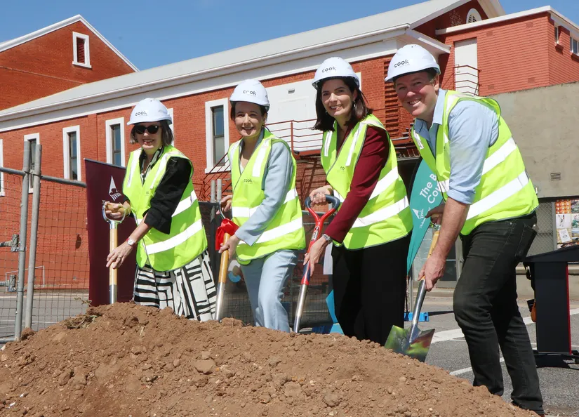 Four people wearing hard hats and holding shovels photographed in the carpark (with original red-brick hall behind), during the official sod turning ceremony.