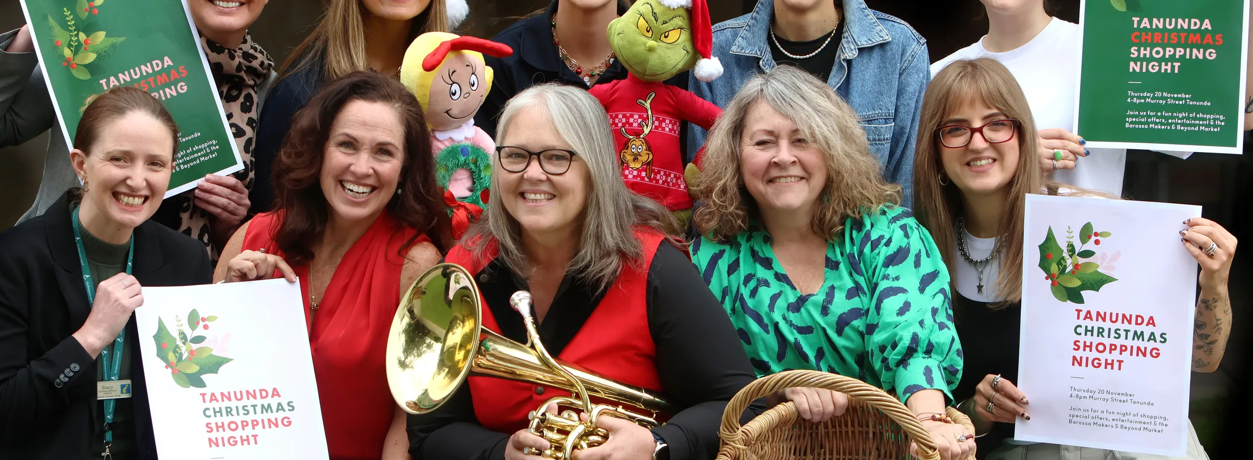 Ten people in a group holding posters, one is holding a brass musical instrument and another with a shopping basket, promoting the upcoming Tanunda Christmas Shopping Night