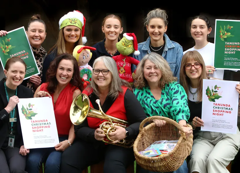 Ten people in a group holding posters, one is holding a brass musical instrument and another with a shopping basket, promoting the upcoming Tanunda Christmas Shopping Night